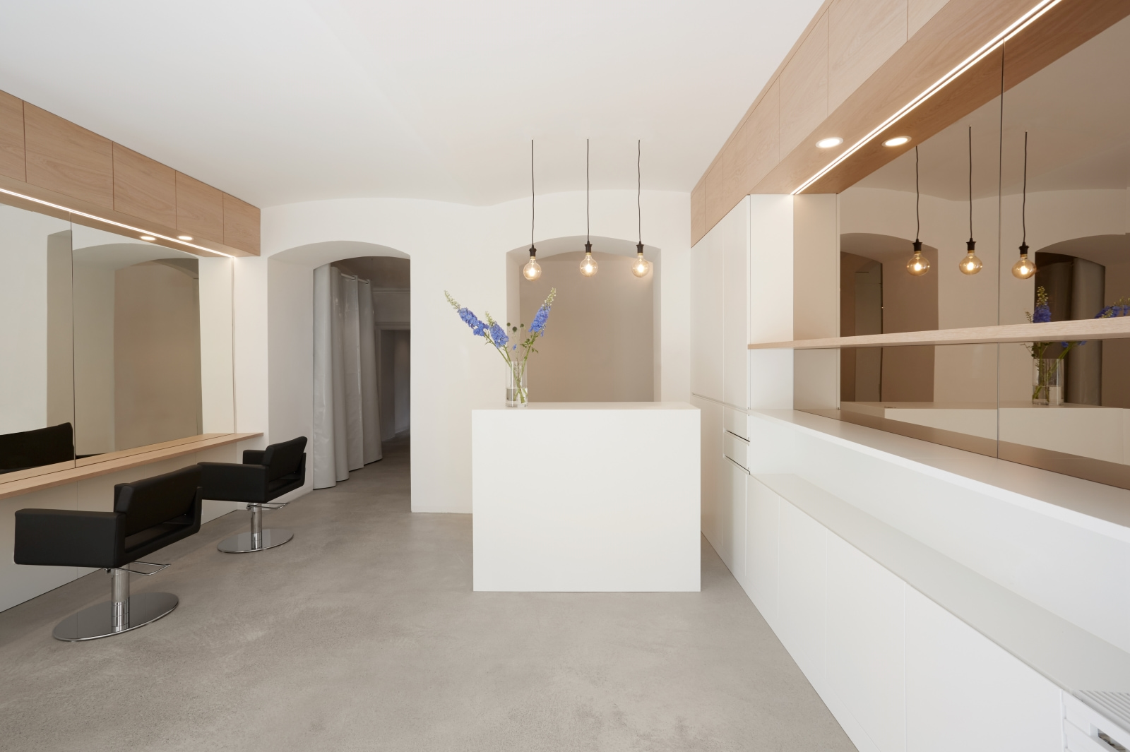 Friseursalon mit weißem Empfangstresen, großen Spiegeln, schwarzen Stylingstühlen und warmen Pendelleuchten im klaren Design.