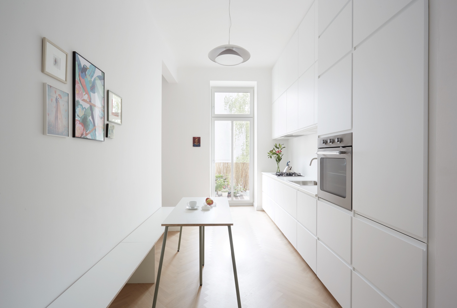 Minimalistische weiße Küche mit durchgehenden Einbaumöbeln, Holzfußboden in Fischgrätmuster, schmalem Esstisch mit Bank und Blick in den grünen Innenhof.