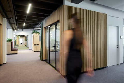 View of the open-plan office with wood-clad room-in-room boxes. Furniture elements and curtains define the circulation and work areas. A person walks through the welcoming space.