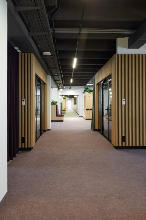 Blickachse durch den hellen, freundlichen Bürogang mit seitlich angeordneten, holzverkleideten Raum-in-Raum-Boxen und lila Akzenten.