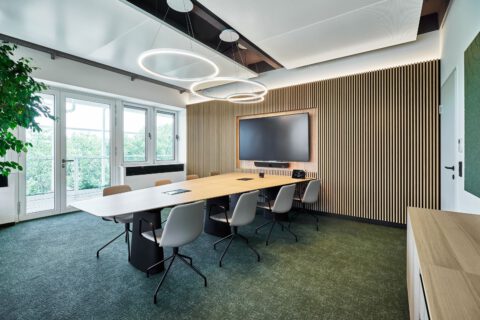 Großer Besprechungsraum mit trapezförmigem Tisch, Wandverkleidung, Bildschirm und Rundhängeleuchte. Pflanzen und Grüntöne prägen die Gestaltung.