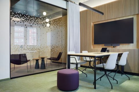 Besprechungsraum mit Wandverkleidung, Bildschirm, Besprechungsstühlen und Pouf. Glaswand mit Vorhang und Folierung, Blick zum Lounge-Wartebereich.