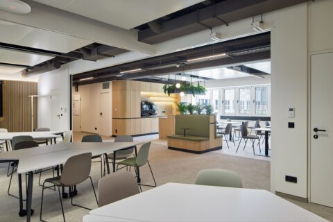 Offener Mehrzweckraum mit Blick auf die Cafeteria. Trapezförmige Tische und Bestuhlung im Vordergrund schaffen eine freundliche, spielerische Atmosphäre.
