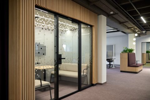 Raum-in-Raum-Box mit Holzlamellen, Glaswand mit CI-inspirierter Folierung und integrierten Sitzbänken für konzentriertes Arbeiten oder Austausch.