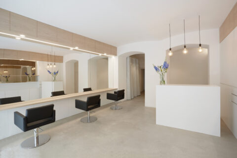 Moderner Friseursalon mit langen Spiegeln, schwarzen Stylingstühlen, weißem Empfangstresen und minimalistischer Raumgestaltung mit klaren Linien.