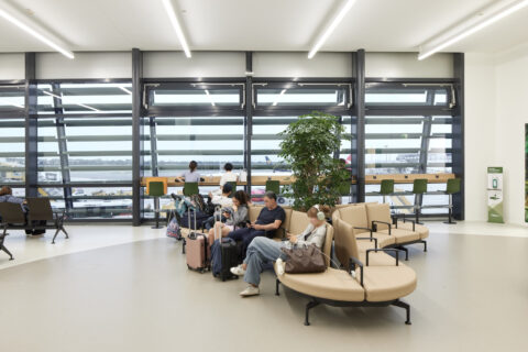Sitzgruppen und Arbeitsplätze mit Panoramafenstern und Ausblick auf das Rollfeld in der Besucherwartezone des Flughafens Wien.