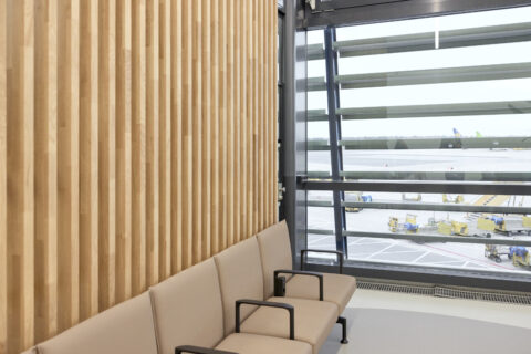 Sitzreihe in der Besucherwartezone des Pier Ost mit eleganter Holzlamellenwand und großem Panoramafenster mit Blick aufs Rollfeld.