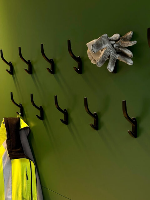 Grüne Garderobenwand mit schwarzen Haken, Schutzkleidung und Arbeitshandschuhen – funktionale Ergänzung zum Aufenthaltsbereich.