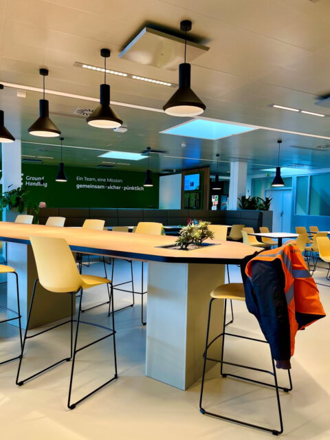 Moderner Aufenthaltsbereich mit großem Gemeinschaftstisch, gelben Barhockern und schwarzen Pendelleuchten. Im Hintergrund grüne Wandgestaltung mit dem Motto ‚Ein Team, eine Mission: gemeinsam – sicher – pünktlich‘.