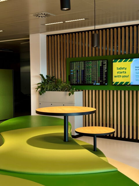 Grüne Sitzlandschaft mit runden Tischen vor Holzlamellen und Monitoren – modernes Design im Aufenthaltsraum.