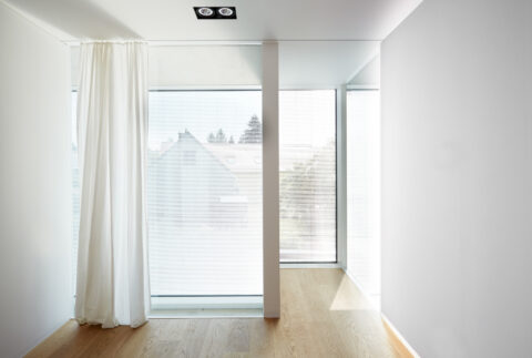 Helles Zimmer mit raumhohen Glasfronten, Vorhängen und Parkettboden. Reduzierte, lichtdurchflutete Architektur mit klarer Formsprache.