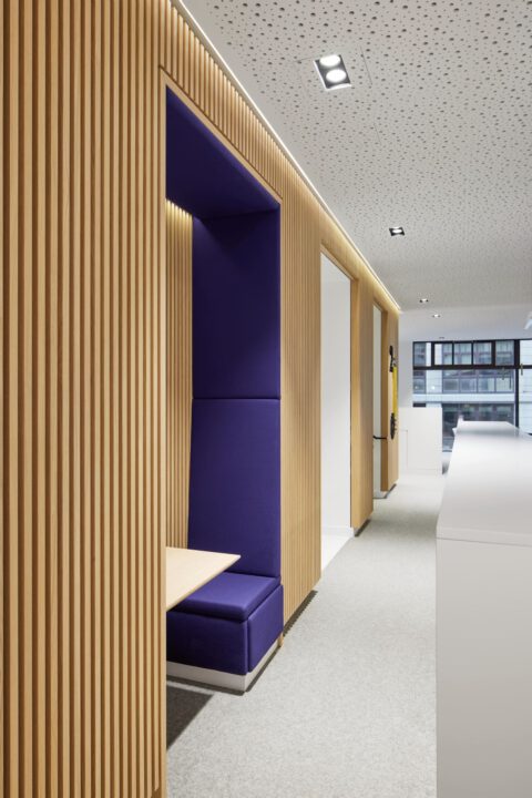 Sitznische mit blauer Polsterung in einer Holzlamellenwand, integriert in einen modernen Büroflur mit hellem Teppichboden und Deckenbeleuchtung.