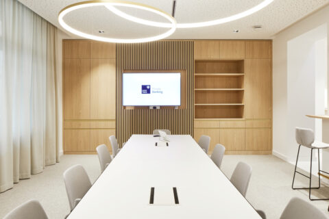 Moderner Besprechungsraum mit hellem Konferenztisch, Holzverkleidung, Designleuchten und Bildschirm für Präsentationen im Private Banking.
