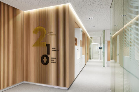 Moderner Büroflur mit Holzlamellenwand und großen Zahlen zur Geschossmarkierung, Glaswänden, heller Deckenbeleuchtung sowie einer kleinen Teeküche/Servicestation.