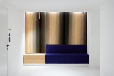 Minimalistischer Wartebereich mit Holzlamellenwand, integrierter Sitzbank in Dunkelblau und modernen Pendelleuchten im klaren Design.