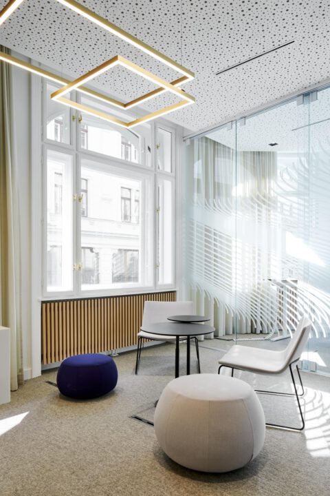 Helles Lounge-Areal mit Glaswand, Designleuchten, Sitzpoufs und klarer Architektur – moderne Raumgestaltung für Beratung und Entspannung.