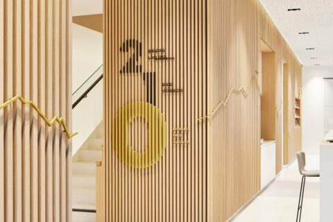 Empfangsbereich eines Bankgebäudes mit vertikaler Holzlamellenwand, großflächigen Geschosszahlen in Gelb und Schwarz sowie eine vergoldete 360-Grad-Kontur der Tiroler Bergkette.