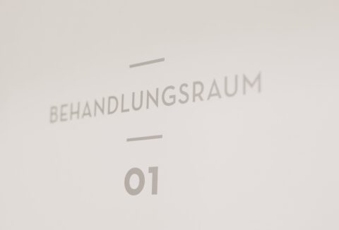 Türbeschriftung mit der Aufschrift „Behandlungsraum 01“ in minimalistischer Typografie.