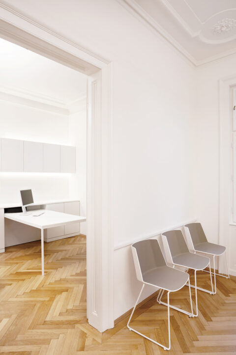 Blick vom Eingang in das Behandlungszimmer mit weißen Wänden, Parkettboden und minimalistischen Stühlen.