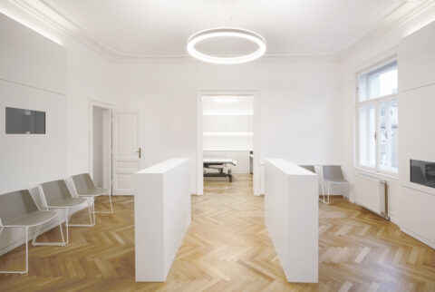 Zentraler Wartebereich mit zwei weißen Empfangstheken, ringförmiger Deckenleuchte und Sitzgelegenheiten.