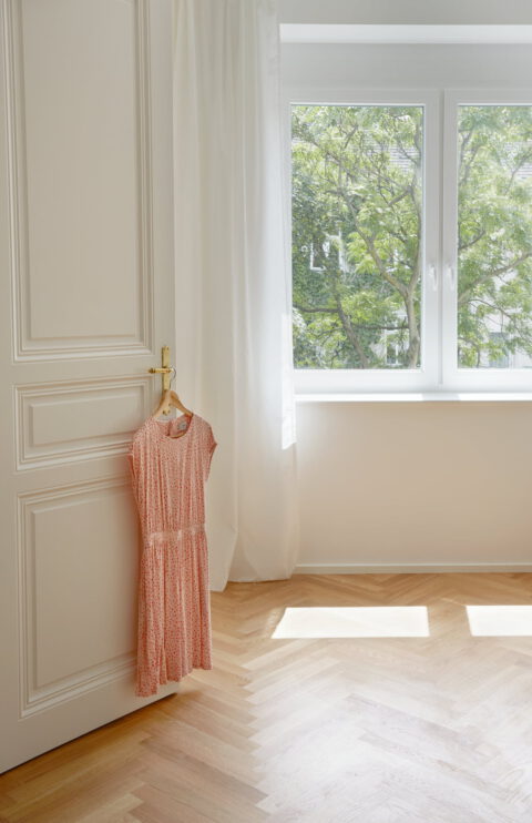 Helles Zimmer mit Fischgrätparkett, weißen Wänden und einem rosa Kleid an der Tür.