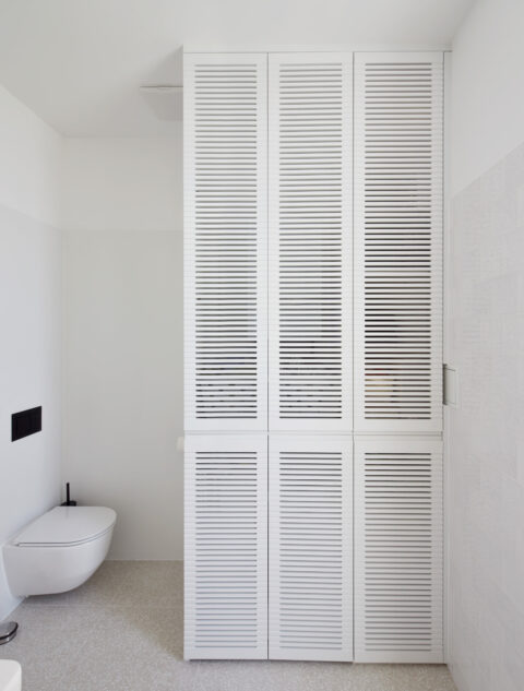 Weißes Badezimmer mit Toilette und hohen Lamellentüren als Schrankverkleidung
