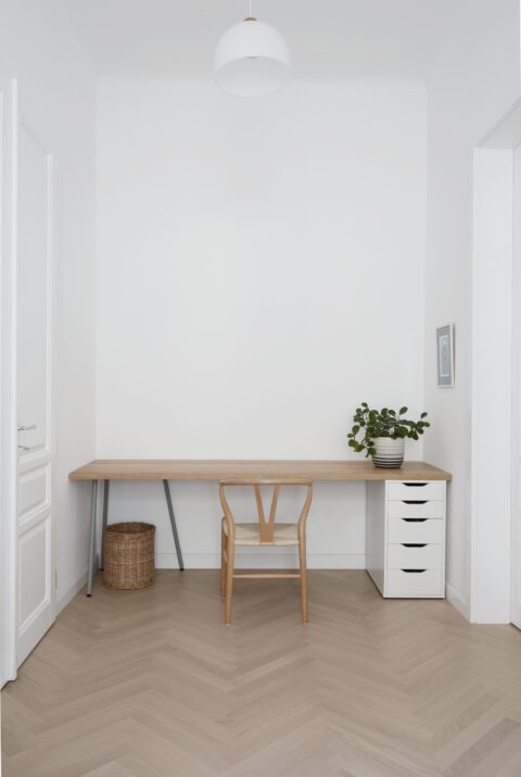 Minimalistischer Arbeitsplatz mit hellem Holzschreibtisch, schlichter Holzstuhl und weißem Schubladenelement, ergänzt durch eine Topfpflanze und Korb, in einem hellen Raum mit Fischgrätparkett.