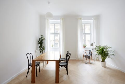 Geräumiges Esszimmer mit großem Holztisch, schwarzen Stühlen, großen Fenstern mit weißen Vorhängen und grünen Zimmerpflanzen.