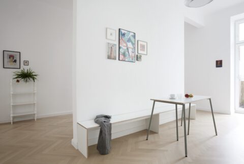 Minimalistischer Essbereich mit kleinem Tisch, eingebauter weißer Bank und gerahmten Bildern an der Wand, neben einem hellen Fenster.