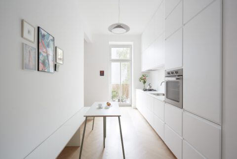Minimalistische weiße Küche mit durchgehenden Einbaumöbeln, Holzfußboden in Fischgrätmuster, schmalem Esstisch mit Bank und Blick in den grünen Innenhof.