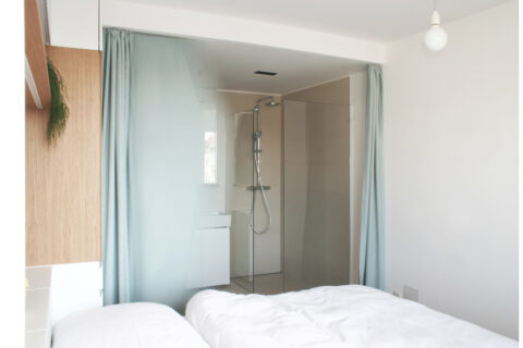 Schlafzimmer mit Blick ins Badezimmer, abgetrennt durch Glaswand und blauen Vorhang.