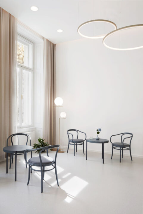 Wartebereich mit hellen Wänden, großen Fenstern, runden Pendelleuchten und klassischen Thonet-Stühlen.