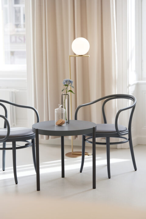 Detailansicht des Wartebereichs mit rundem Tisch, zwei Thonet-Stühlen und einer eleganten Stehleuchte mit Kugelleuchte.