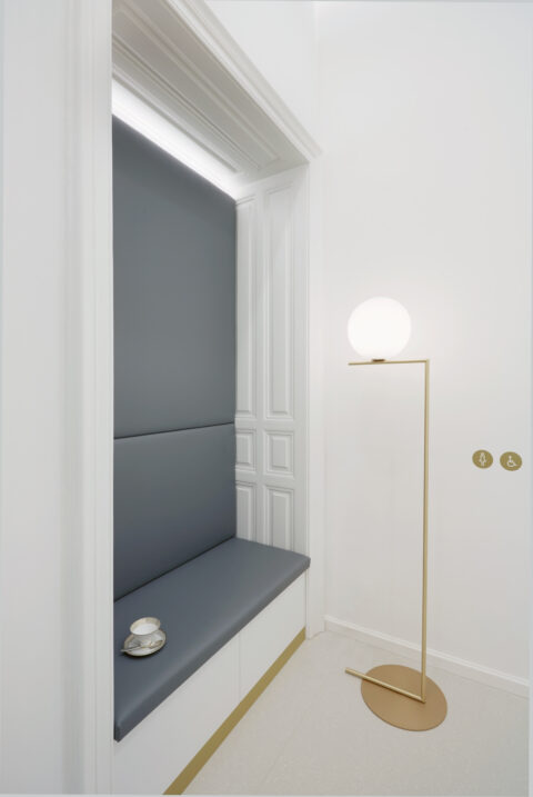 Sitznische mit gepolsterter Bank in graublauem Stoff, flankiert von einer goldenen Stehleuchte mit Kugelleuchte.