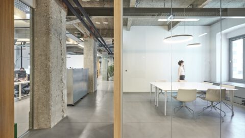 Blick durch Glaswand in einen minimalistischen Besprechungsraum mit weißem Tisch und modernen Stühlen, flankiert von Sichtbetonstützen.